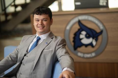 Westfield State student in a gray suit smiles while seated in front of a university podium featuring the Westfield State owl logo.