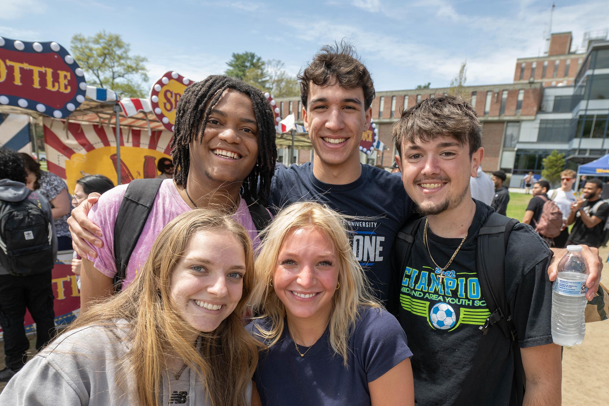 A group of five ,ale and female Westfield State students smile at the camera during the FlightFest carnival on the campus green