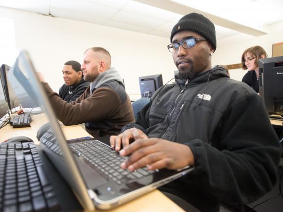 Male student in glasses and beanie cap working on laptop in class