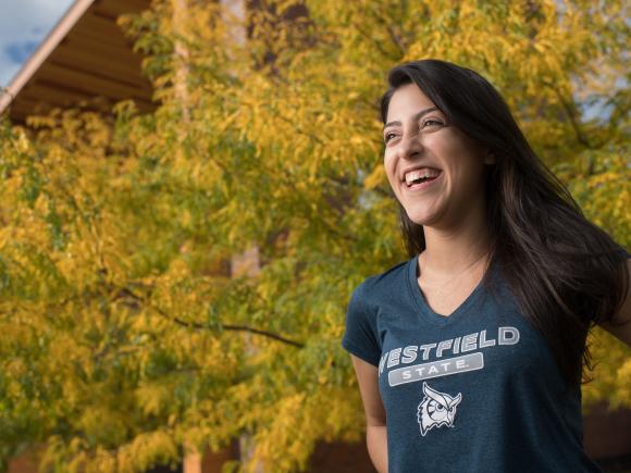 Female student in a Westfield State t-shirt in front of tree with autumn foliage