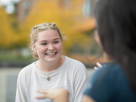 Image of a female student smiling at her friend as they talk on the campus green