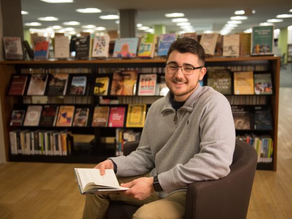 make student in glasses sitting in the Ely Library with a book in his lap