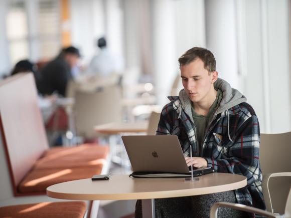 Male student works on his laptop at a table inside the Stevens Center