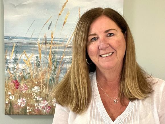 A white woman is standing in front of a beach painting and smiling