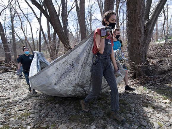 Three students wearing masks carry an open tarp through a nature area.