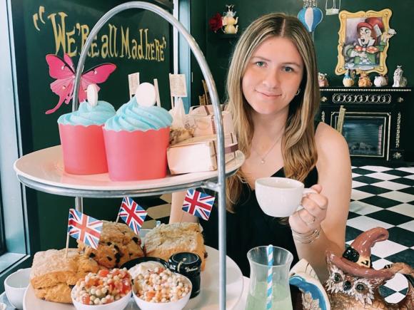 Westfield State student Madeline LeBlanc holds a tea cup and looks out from behind a tray of sandwiches and desserts in a restaurant decorated with an Alice in Wonderland theme.