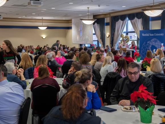 Large crowd of Westfield State employees sit at tables in Scanlon Banquet Hall decorated with poinsettias while President Linda Thompson reads names of raffle winners at the podium.