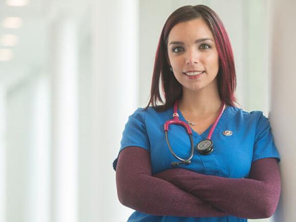 A graduate student wearing blue scrubs and a stethoscope leans against a wall while posing for a photo.