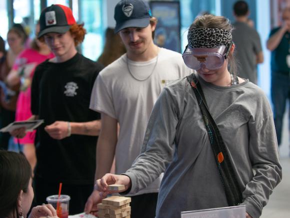 A student with goggles plays Jenga with another student behind him smiling.