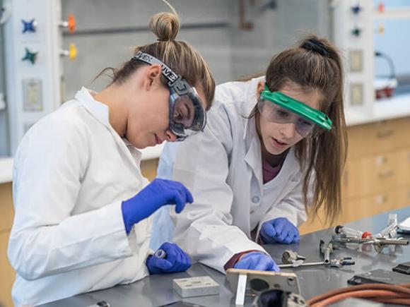 Two students wearing lab coats, gloves, and safety goggles conduct an experiment in a laboratory setting.