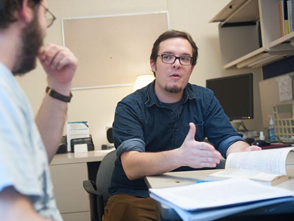 A professor sits in their office and explains something from a book to a student sitting across from them.