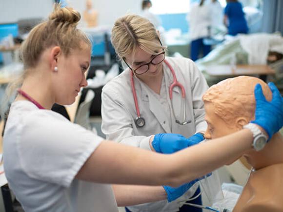 Two students wearing scrubs, stethoscopes, and gloves practice on a medical mannequin.