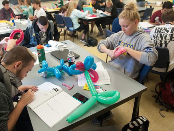 Students sit around large tables covered with balloon animals while working on an assignment.