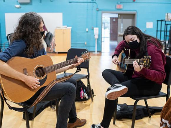 Two students practice playing acoustic guitar in a music room.