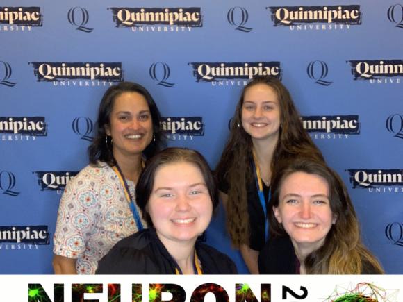 Karina Little ('23), Jamie Gross ('24), and Jamie Goodall ('25), along with Dr. Princy Mennella, smile and pose in front of a blue, Quinnipiac University poster.
