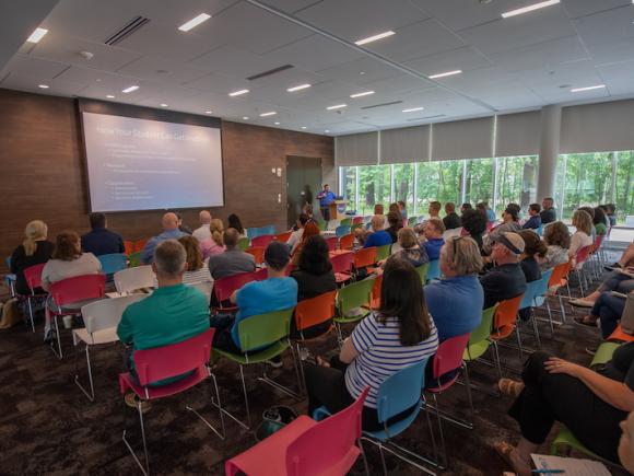 Multi-colored chairs hold new students and their Champions while presentations about new student orientation are given via a projector.