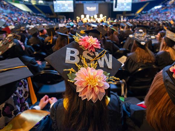  A student seated at a graduation ceremony wears a graduation cap decorated with flowers and the letters BSW.