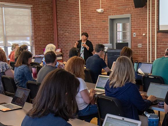 A professor lectures at the front of a classroom while students take notes on their laptops.