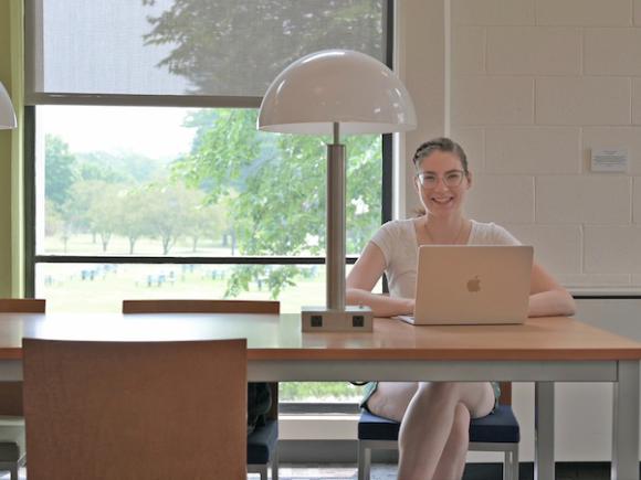 Elise Miller, a white, brown-haired woman with glasses and a ponytail sits behind a desk with a laptop on it.