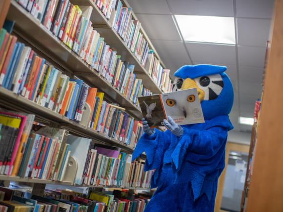 Nestor, a blue colored Owl mascot, reads a book with a white owl face on its cover.