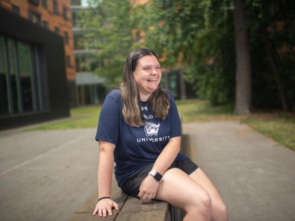 Emily Czupta, a brown-haired, white woman wearing a navy blue T-shirt and black shorts smiles to the side. She sits on a stone bench near a brick building. Green trees dot the background, which is blurred.