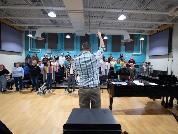A chorus of various WSU students is facing a professor in a blue-plaid button down as he lifts his arms in the air and conducts them.