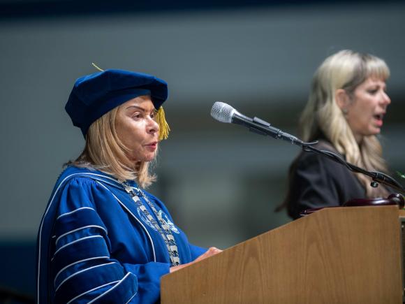 President Linda Thompson stands at the podium during Commencement 2023 wearing blue academic regalia with signer in background