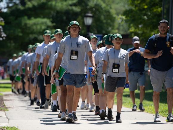 A photo of two lines of people walking in formation, wearing blue shorts, grey shirts, and baseball caps. They also have cadet police lanyards around their neck, as part of the •	The North East Regional Law Enforcement Education Association's camp.