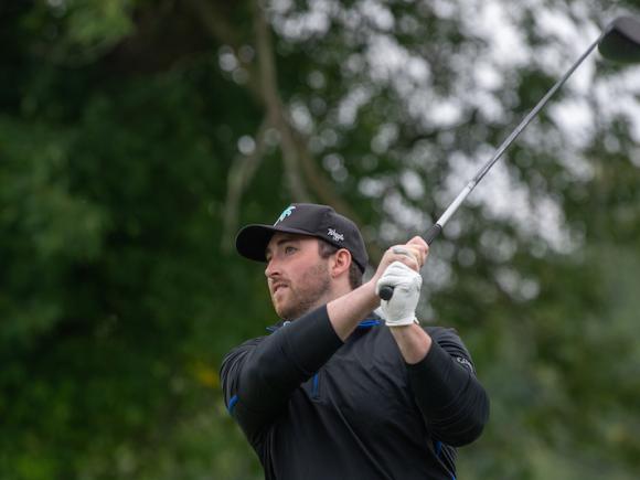 A photo showing a young, white man in a black baseball cap and sweater mid-swing at a golf tournament.