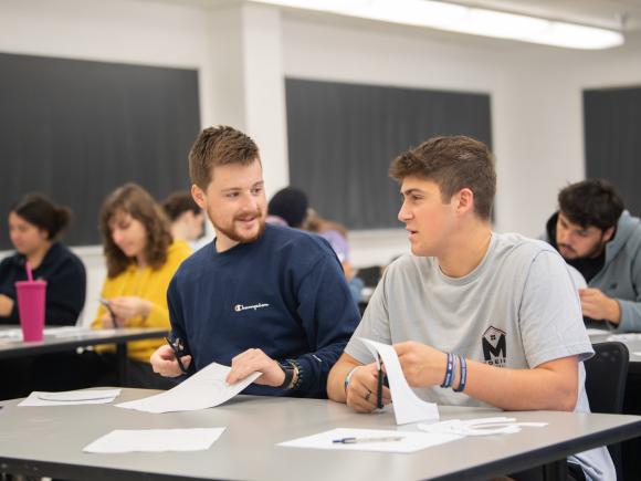 Two students in a classroom talking holding paper assignments