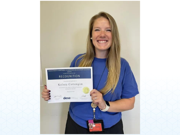 Kelsey Cottengim, an alumna of WSU, wears a navy blue T-shirt and holds a paper that says, "Certificate of Recognition" for the Massachusetts Distinguished Educator Award for 2023.