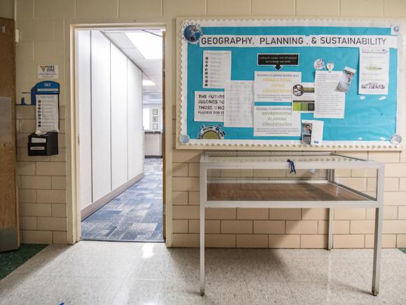 A blue bulletin board with "The Geography, Planning, and Sustainability Department" written across it. White posters and pieces of paper decorate the bulletin board, which rests against white, tiled walls.