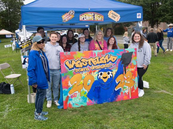 President Linda Thompson and a group of students pose behind a colorful poster of Nestor for Homecoming. The blue tent behind them says, "Lights! Cameras! Homecoming!"