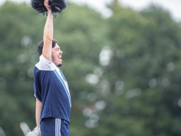 A male cheerleader dressed in navy blue throws one hand into the air while he cheers.