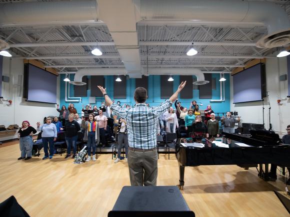 Music class with instructor with hands raised in front of piano with a class of students.