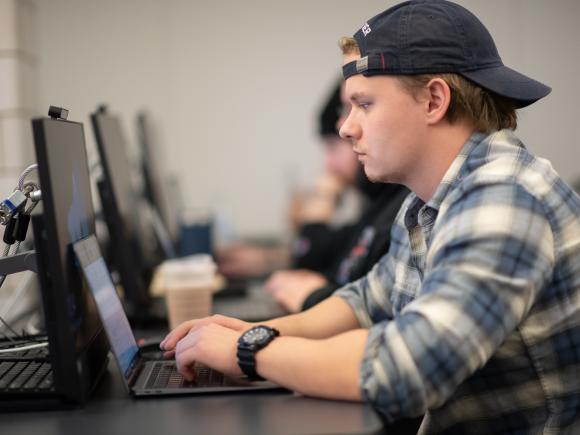 Data science student at desk top computer wearing plaid shirt and baseball hat