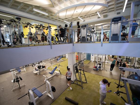 A shot of the Ely Fitness Center, shot from halfway on the staircase leading up and down. White cardio machines are stacked in a row on the upstairs portion of the room with white benches down below for weight lifting.