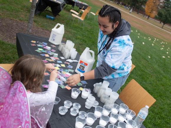 A student in a tie-die sweater makes arts and crafts outside with a young girl in a butterfly costume at a table.