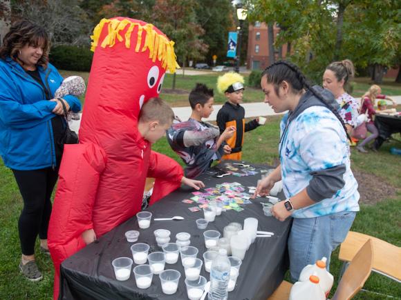 A student in a tie-dye sweater does arts and crafts with a young child in a red monster costume with several other dressed up children around him.
