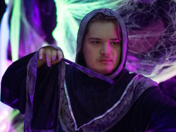 A student in a black robe holds a magic wands and is surrounded by fake spider webs and neon, purple light.