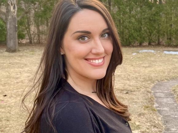 Desiree Nickel '11, a woman with long, brown hair, smiles with pine trees from an unnamed forest in the background. 