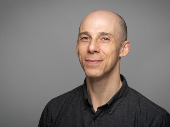 Man smiling wearing a dark grey shirt against a grey background.