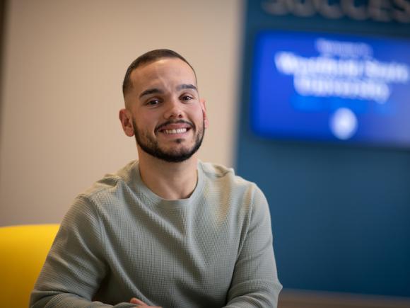 Nate Ferreira, sitting on a yellow chair with a blue and white background behind him that is out of focus.