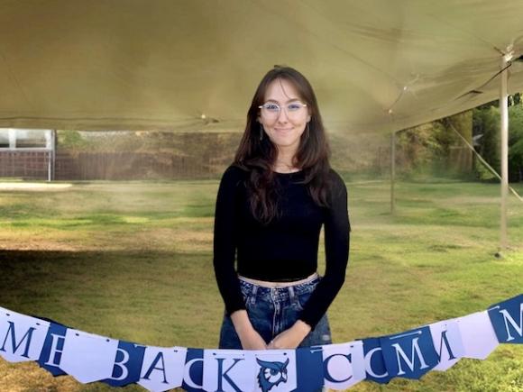 Samantha Palmer '24, in a long, black-sleeve shirt and jeans. She wears glasses and stands before a blue and white banner that says, "Welcome Back Comm Majors."