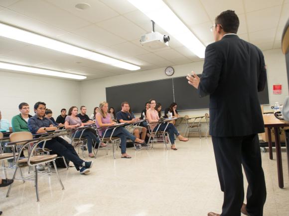 Accounting class with instructor standing in front of room of students wearing a suit.