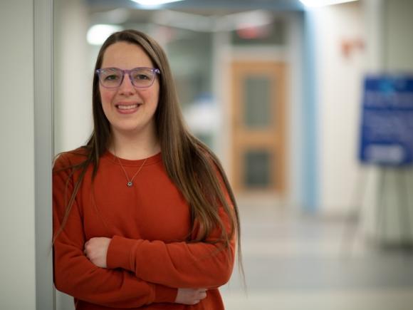 Chelsea Baker, class of 2015, leans against a white doorway. She is wearing an orange long-sleeved shirt and glasses. She's smiling directly at the camera while an out-of-focus hallway is in the background.