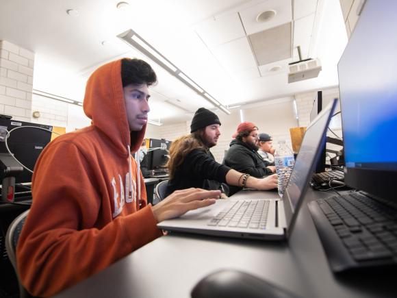 CIS student wearing orange hoodie working on laptop in classroom.