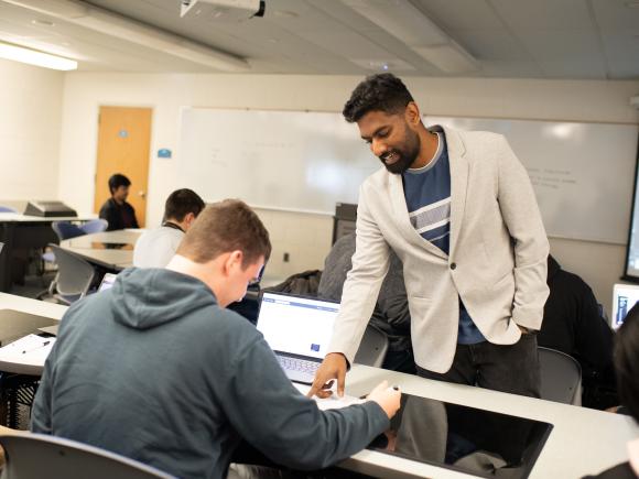 Computer science faculty member wearing white coat assisting a student wearing navy hoodie.