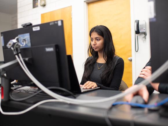 Data science student with long dark hair working in front of a desktop computer in classroom.