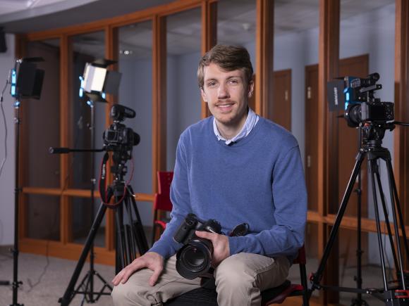 Nathan Dion, class of 2023, sits among a three professional cameras that he uses at John Garvey Inc., as their new Digital Public Relations Analyst. He wears a long-sleeved, blue sweater and tan pants. He holds a black camera in his lap while posing on a stool.